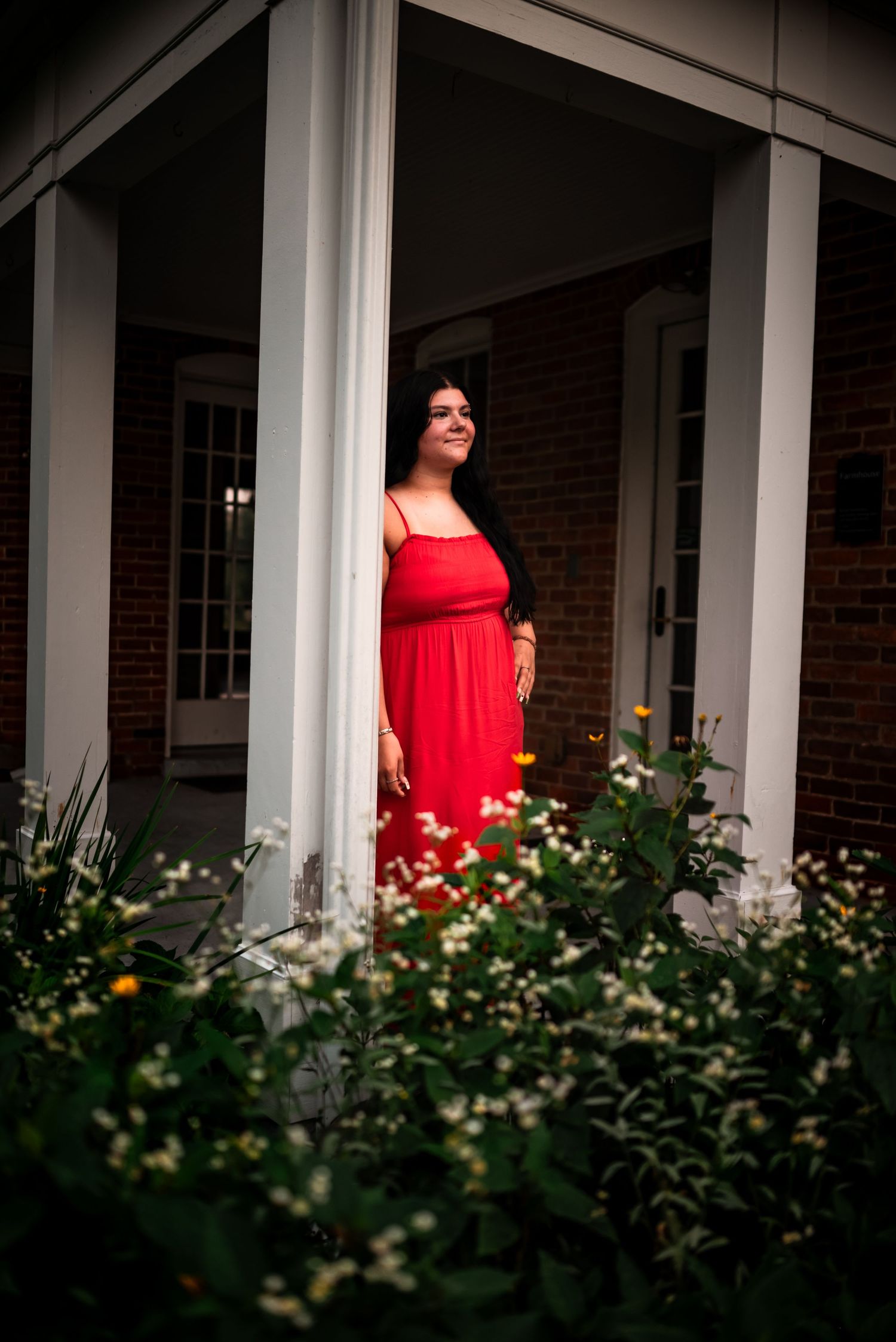 A figure in a vibrant red dress stands on a white-columned porch with lush greenery in the foreground.