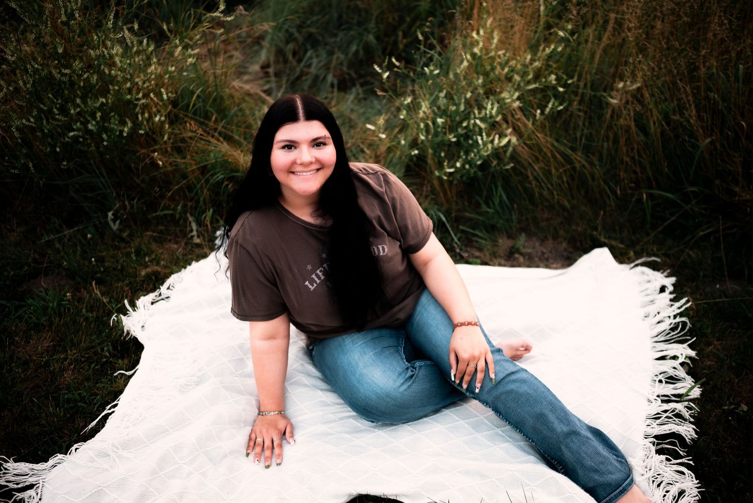 A person in a brown t-shirt and jeans sits on a white blanket outdoors against a backdrop of greenery.