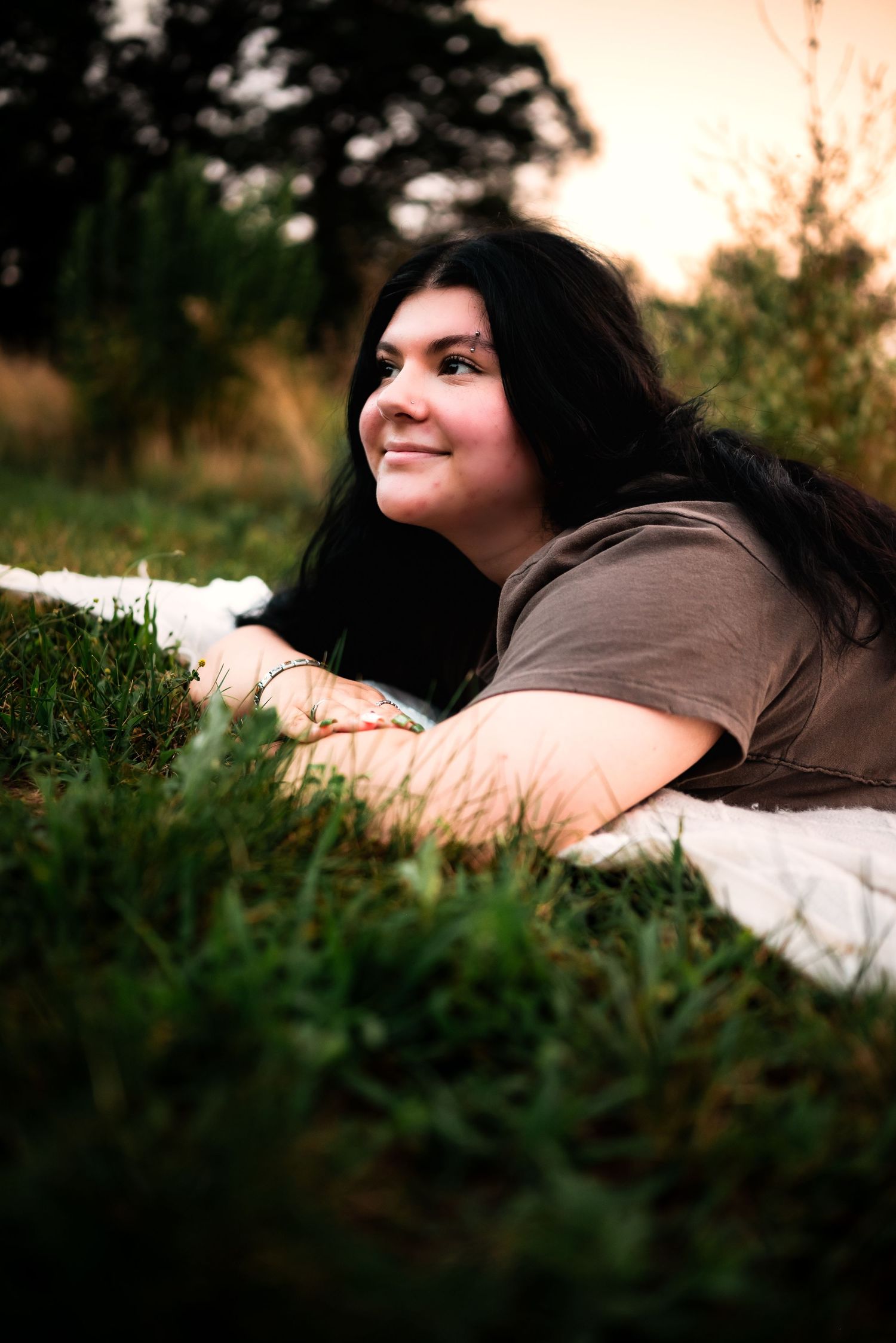 Person with long dark hair lying on a blanket in green grass during golden hour, looking up with a smile.