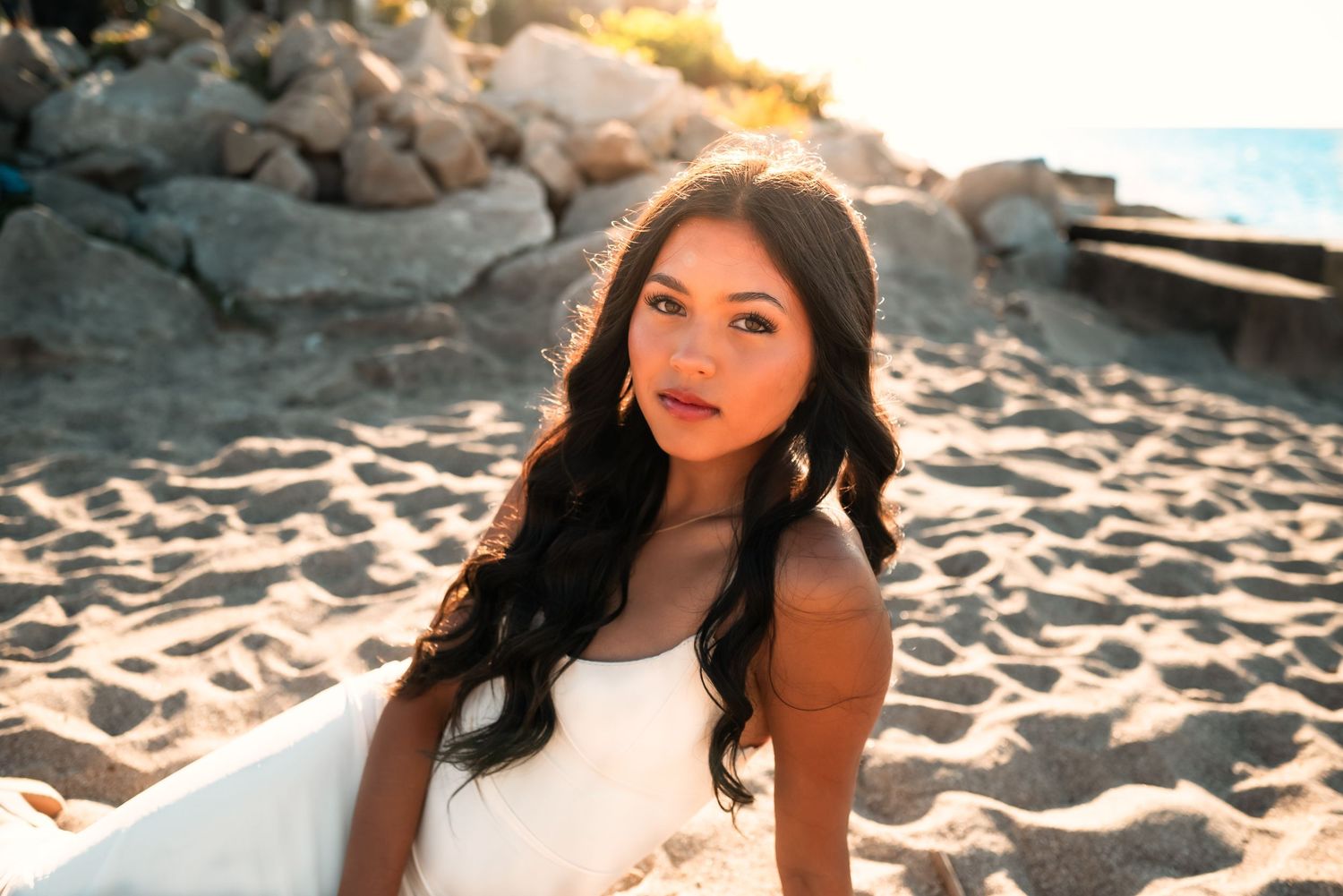 A person with long dark hair poses on a sandy beach in a white dress during golden hour lighting.