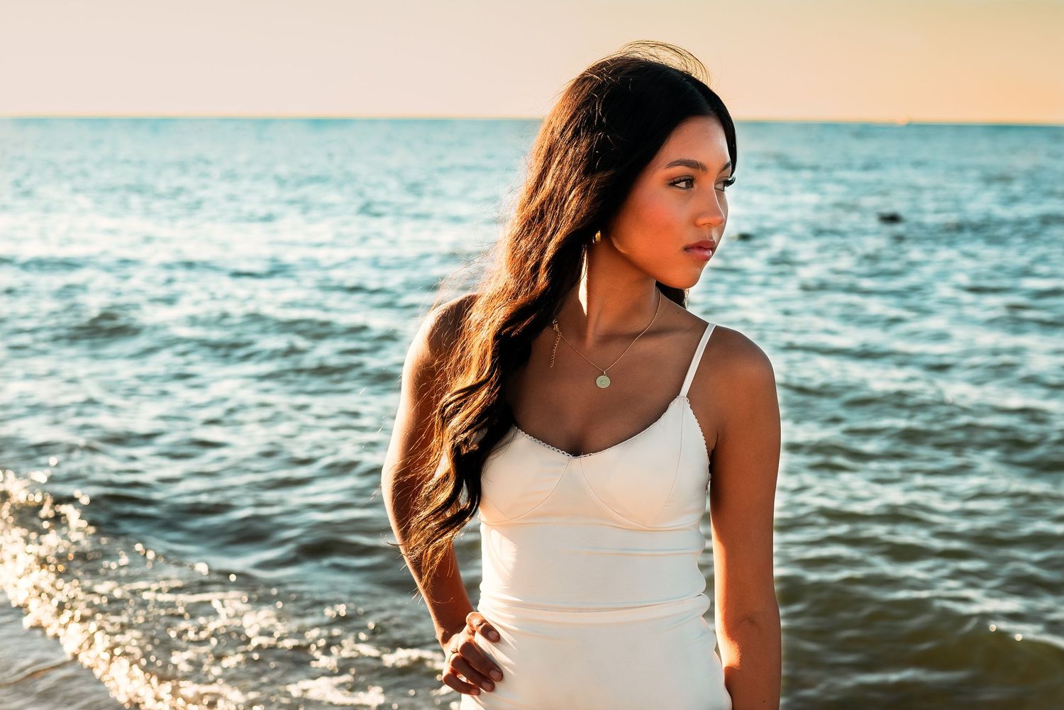 A person in a white dress poses at the beach during sunset with ocean waves in the background.