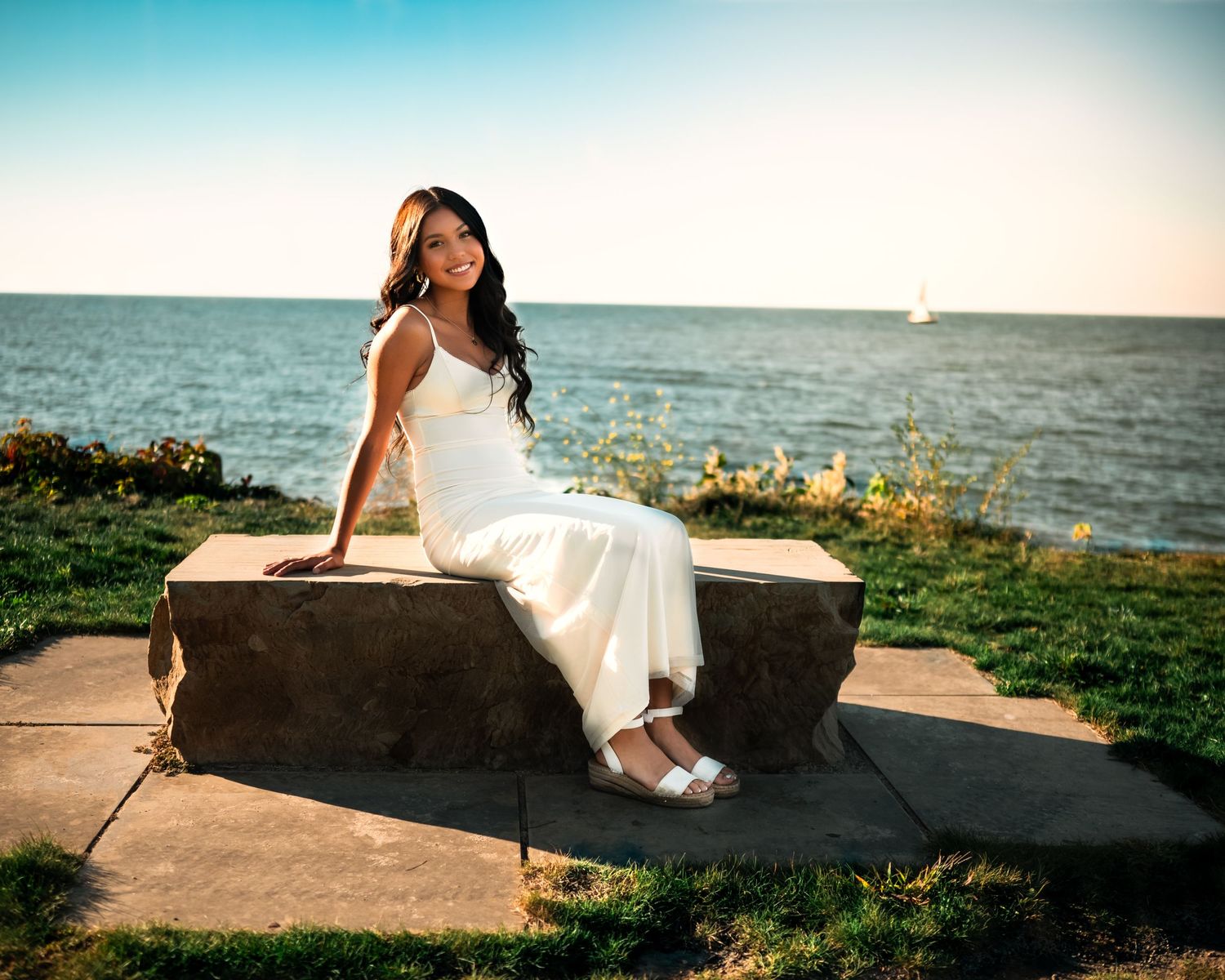 A person in a white dress sits on a stone wall by the ocean during golden hour sunset.