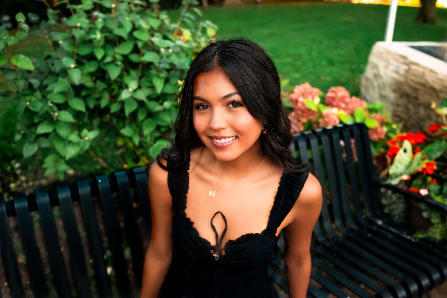 Someone in a black dress sits on a garden bench surrounded by colorful flowers and greenery on a sunny day.
