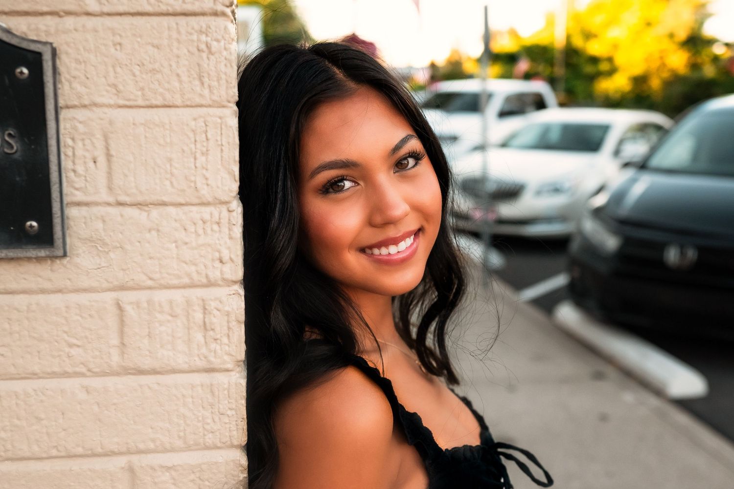 Woman with long dark hair smiling while posing outdoors against a beige wall and parked cars.