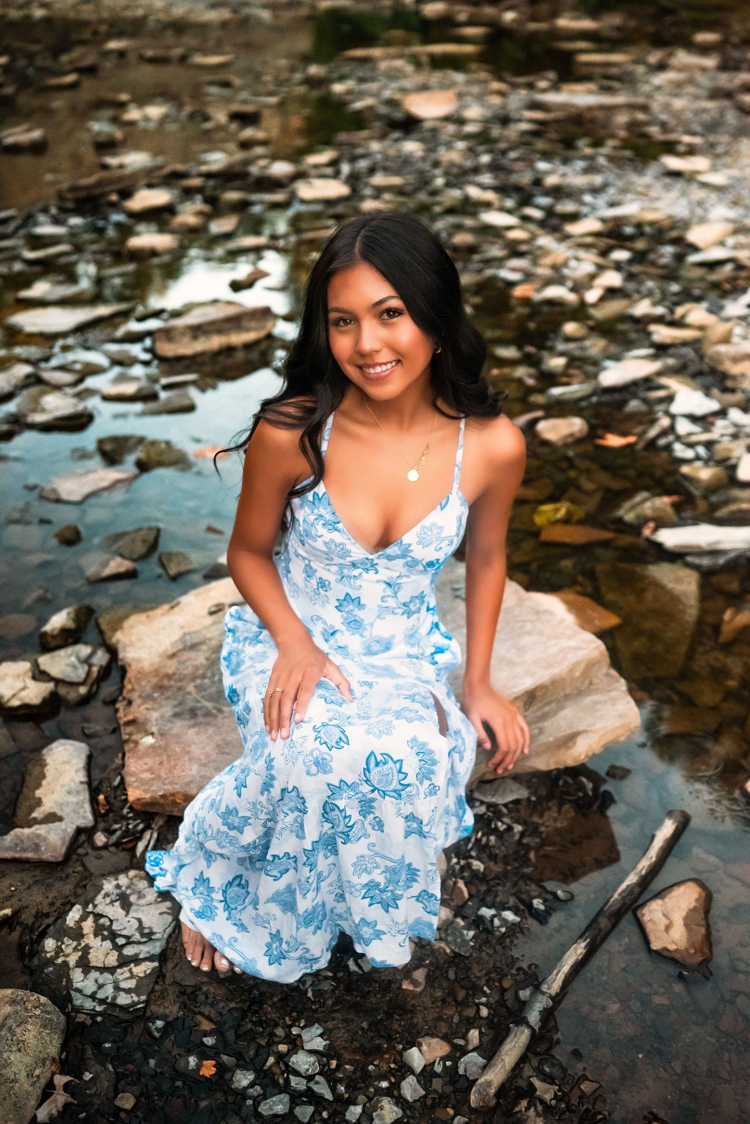 Woman in blue and white floral dress poses gracefully by a rocky stream during an outdoor photo session.