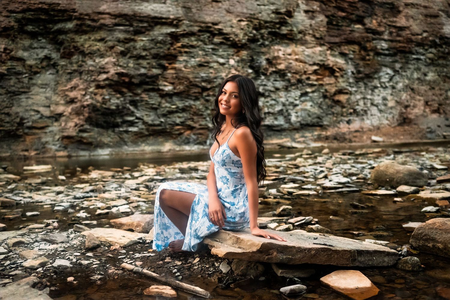 A person in a floral dress poses gracefully by a rocky stream in a natural outdoor setting.