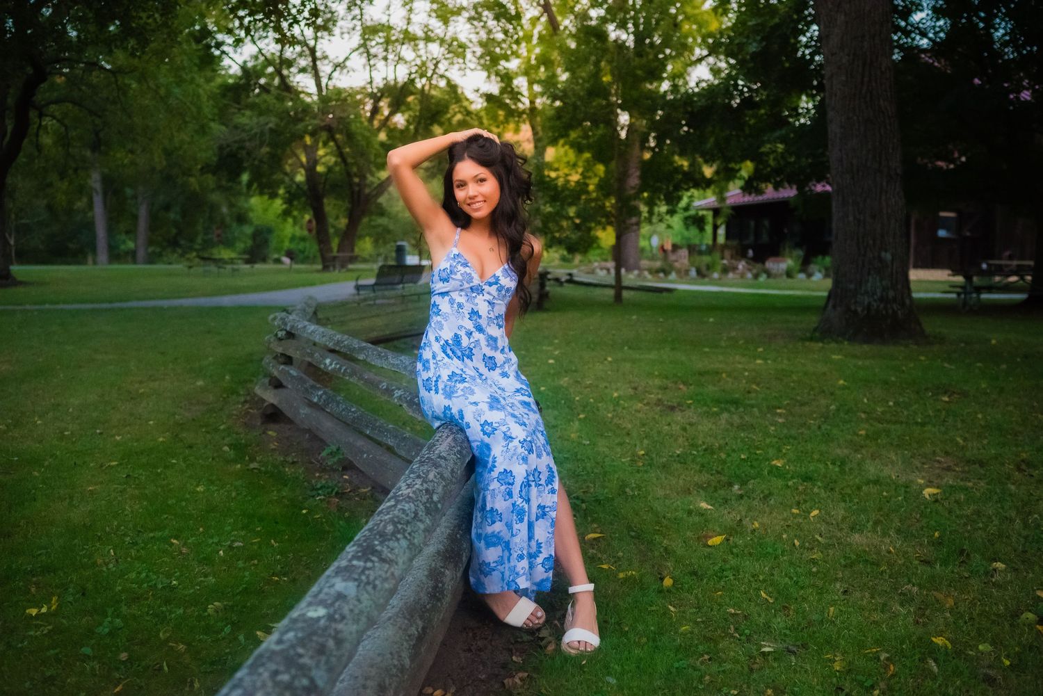 A person in a blue and white floral dress poses playfully on a stone wall in a lush green park setting.