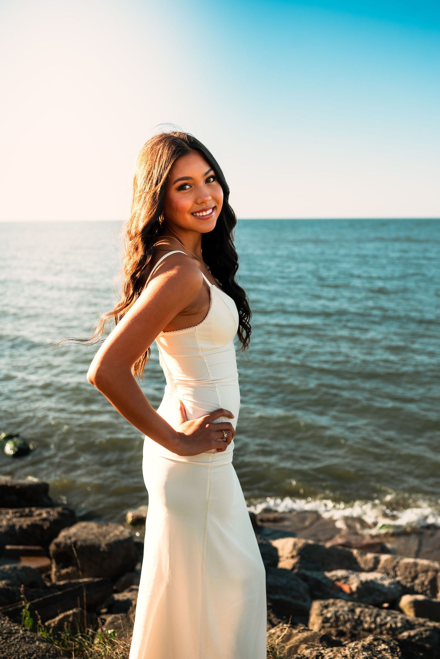 A person in a white dress poses along a rocky coastline with blue ocean and sky in the background.