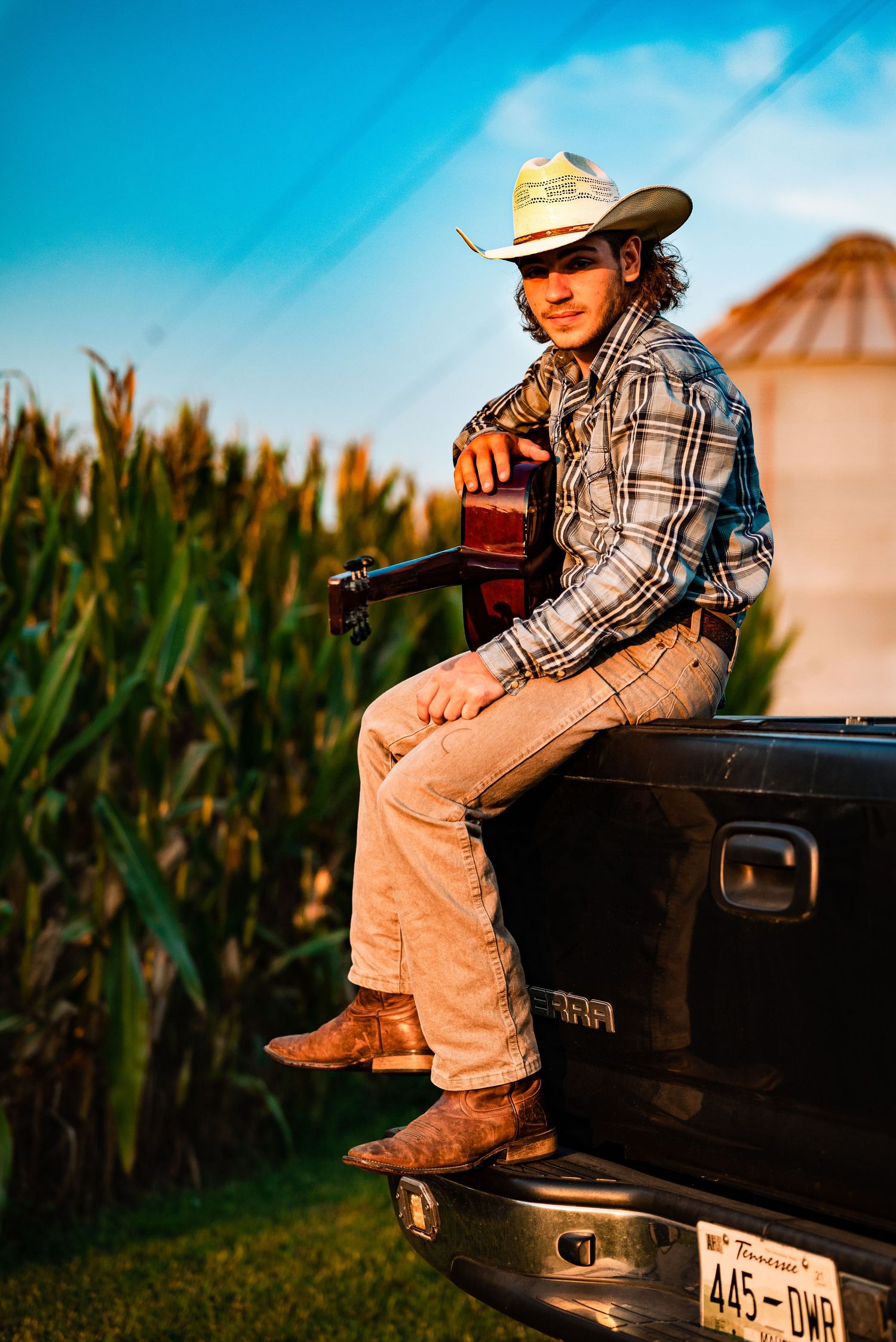 Person in western wear sitting on black truck tailgate near cornfield at sunset.