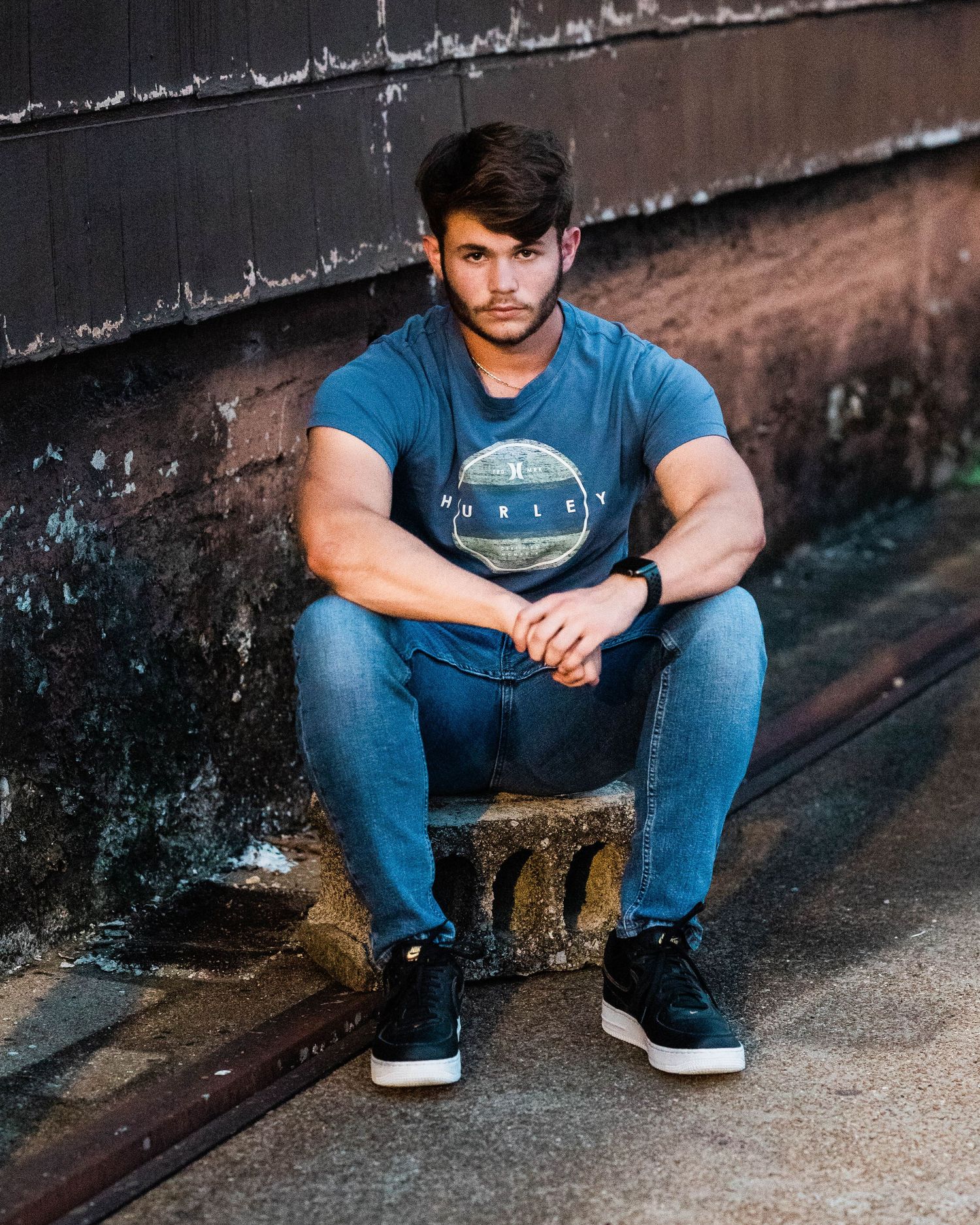 Casual street photography showing person in denim and t-shirt sitting against urban wall.