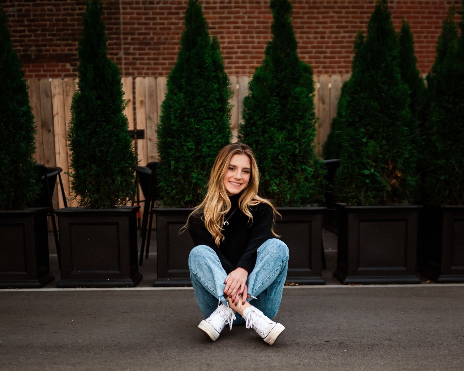 Urban fashion shoot with decorative evergreen trees and brick wall backdrop.