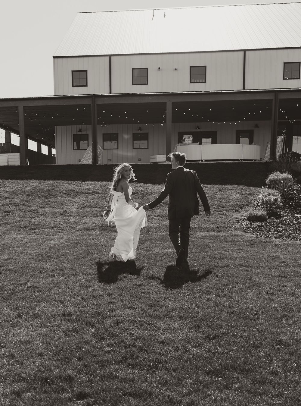 A bride and groom walk hand in hand away from a rustic venue in black and white.