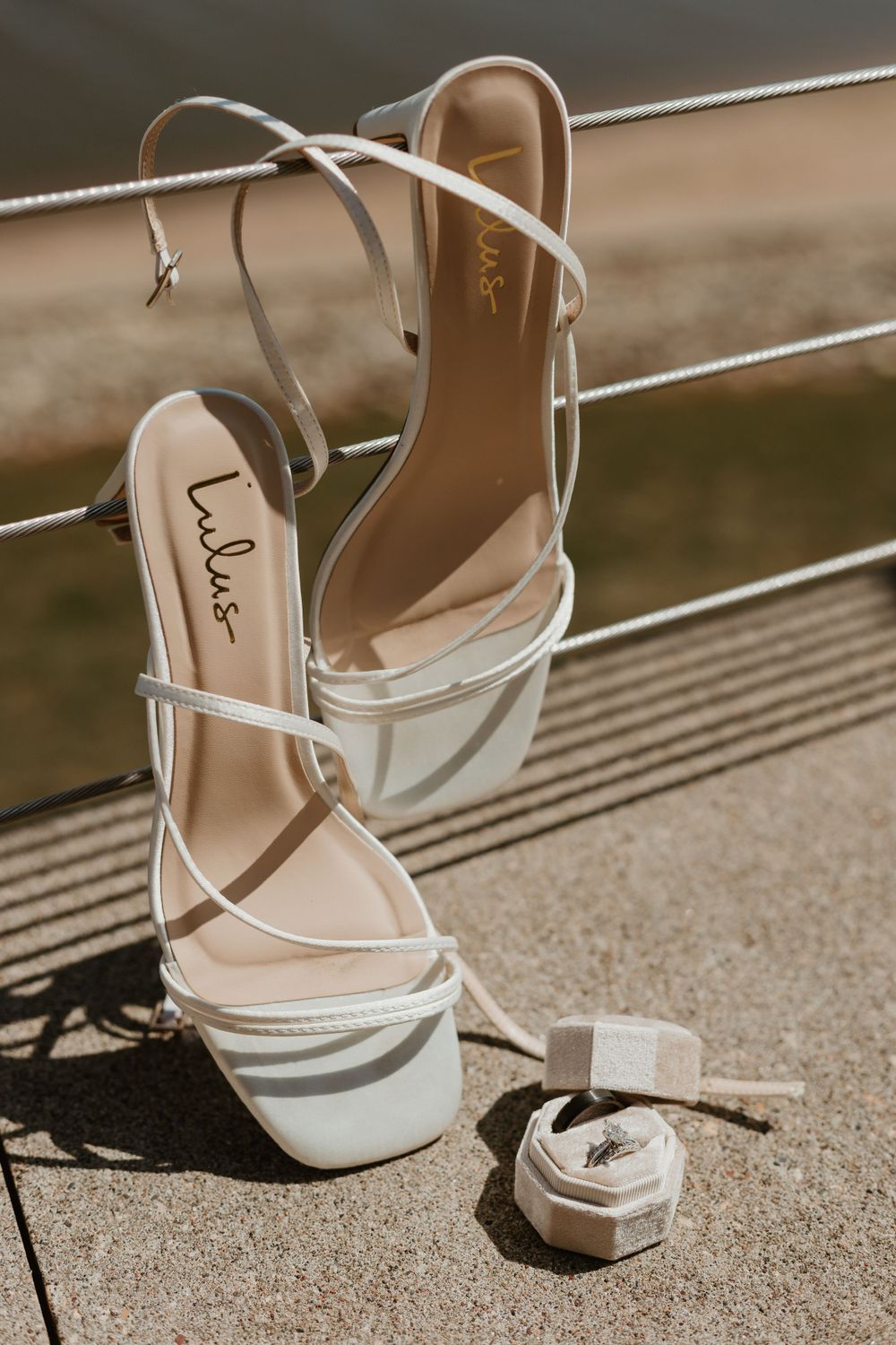 Elegant white strappy wedding heels arranged on metal railing with sunlight.