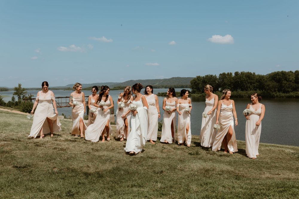 A group wearing champagne dresses poses together on a grassy hill overlooking a lake.