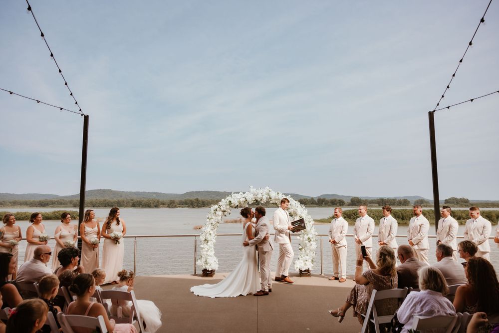 An outdoor waterfront wedding ceremony with string lights and guests seated on white chairs.