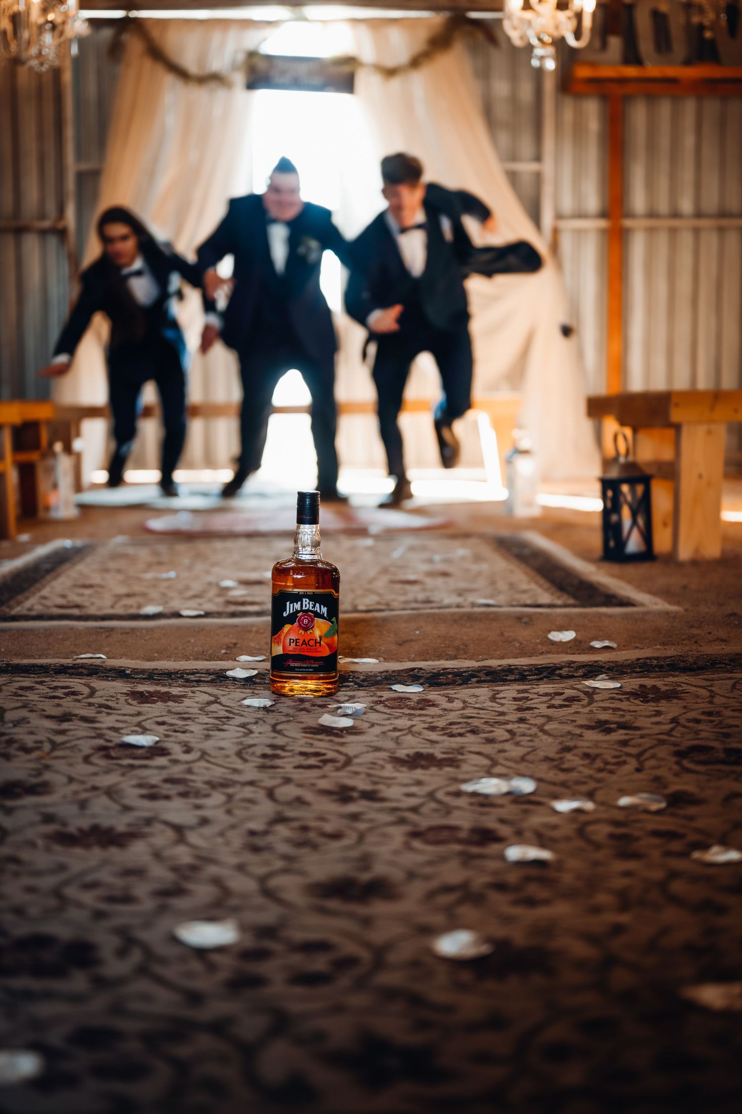 Groomsmen jumping joyfully in a barn wedding venue with a Jim Beam whiskey bottle in the foreground.