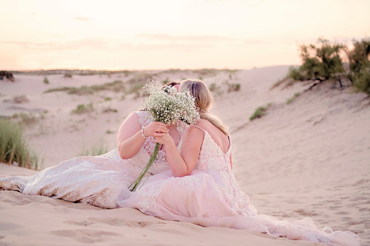 Two brides in pink gowns sit on sandy dunes at sunset, sharing a bouquet of white wildflowers.
