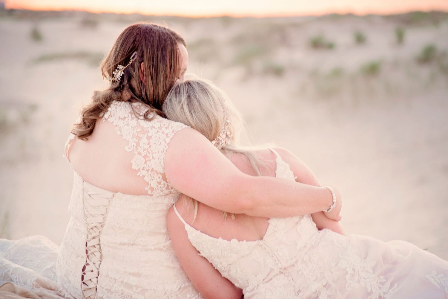 Two brides in lace wedding gowns embrace on a sandy beach at sunset, sharing a romantic moment.