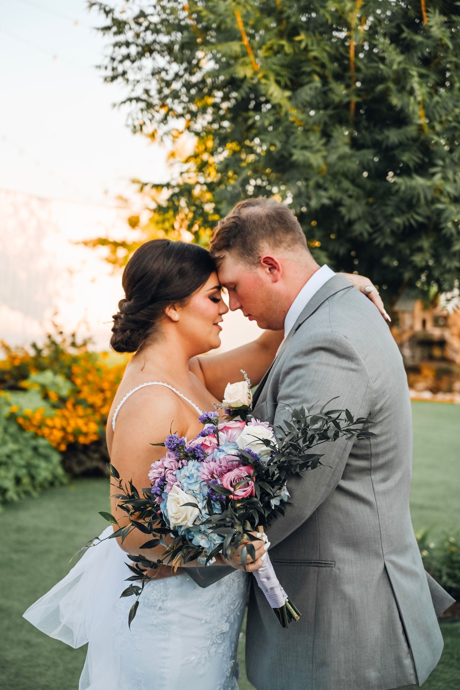 Bride and groom touching foreheads tenderly outdoors, bride holding colorful bouquet of pink and blue flowers at golden hour.