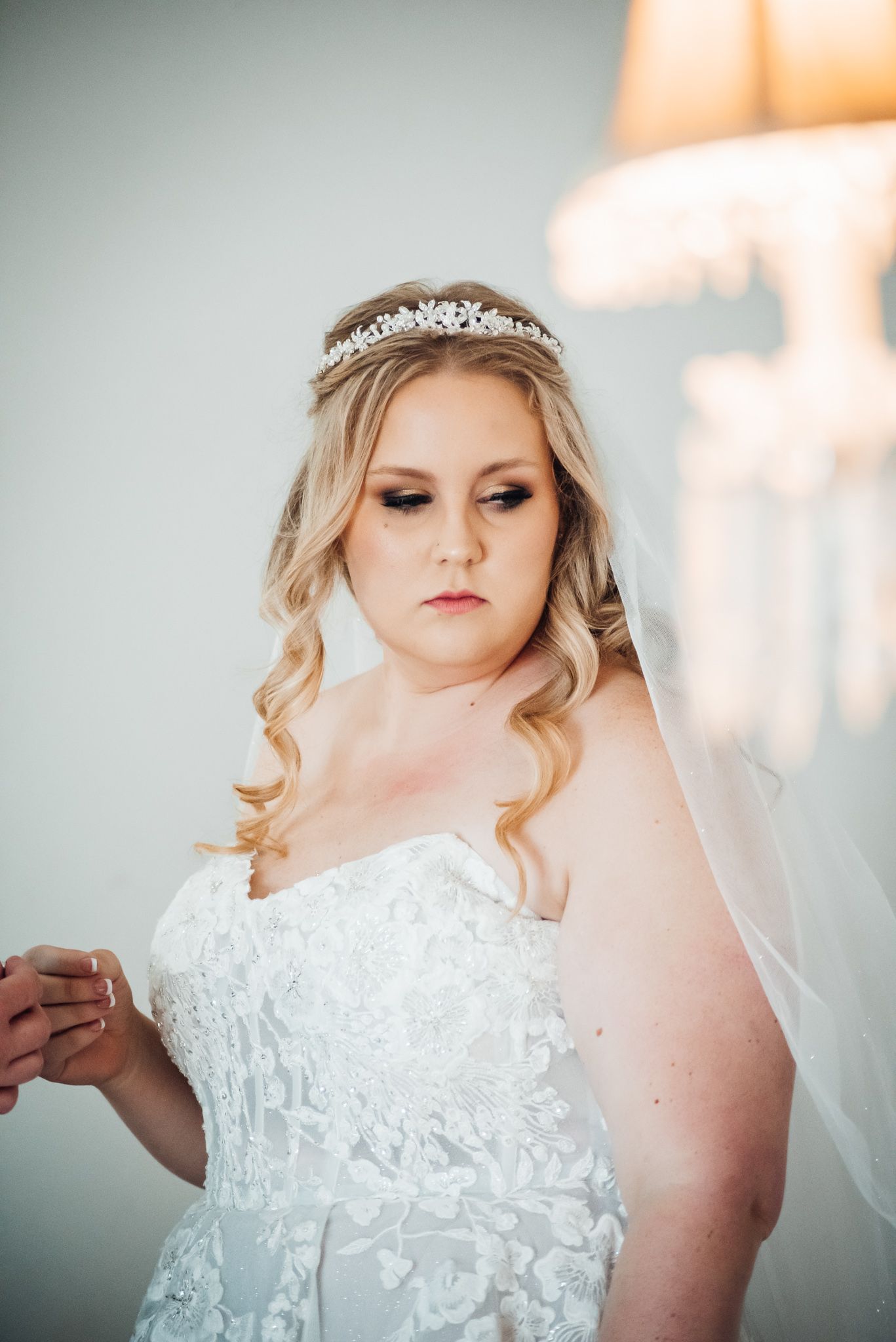 Blonde bride in strapless lace gown with tiara and veil looks down elegantly.