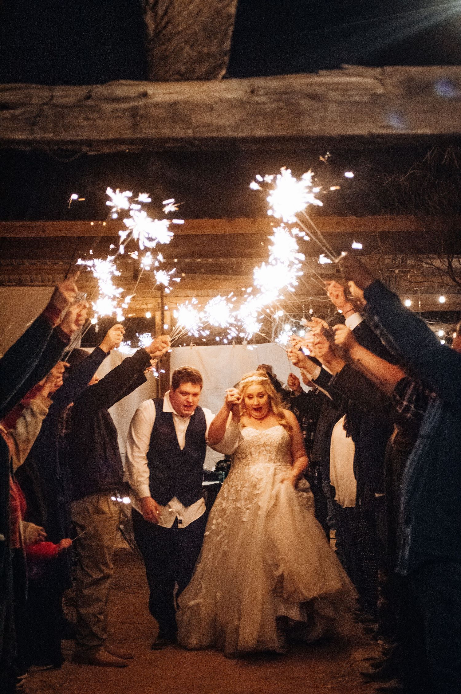 Bride and groom exit through sparkler tunnel at rustic barn wedding reception at night.