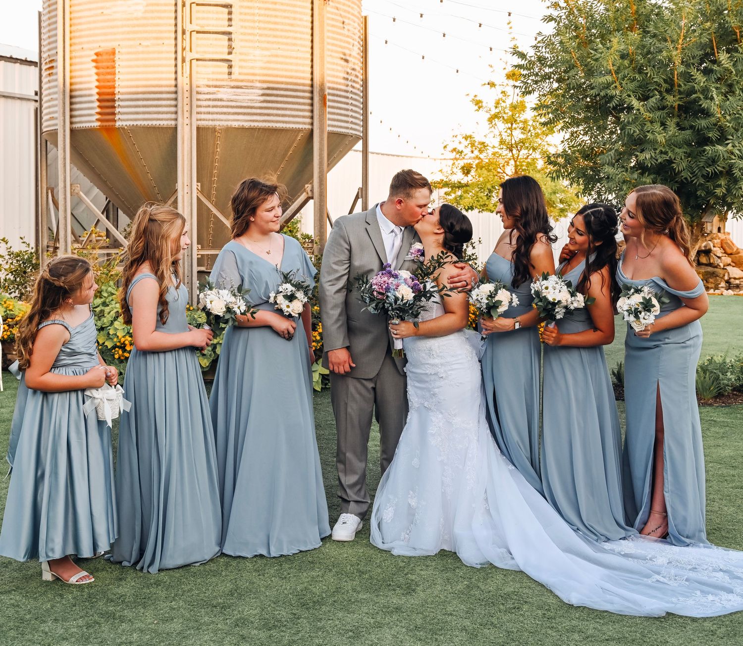 Wedding couple kissing surrounded by bridesmaids in dusty blue gowns holding bouquets at rustic farm venue.