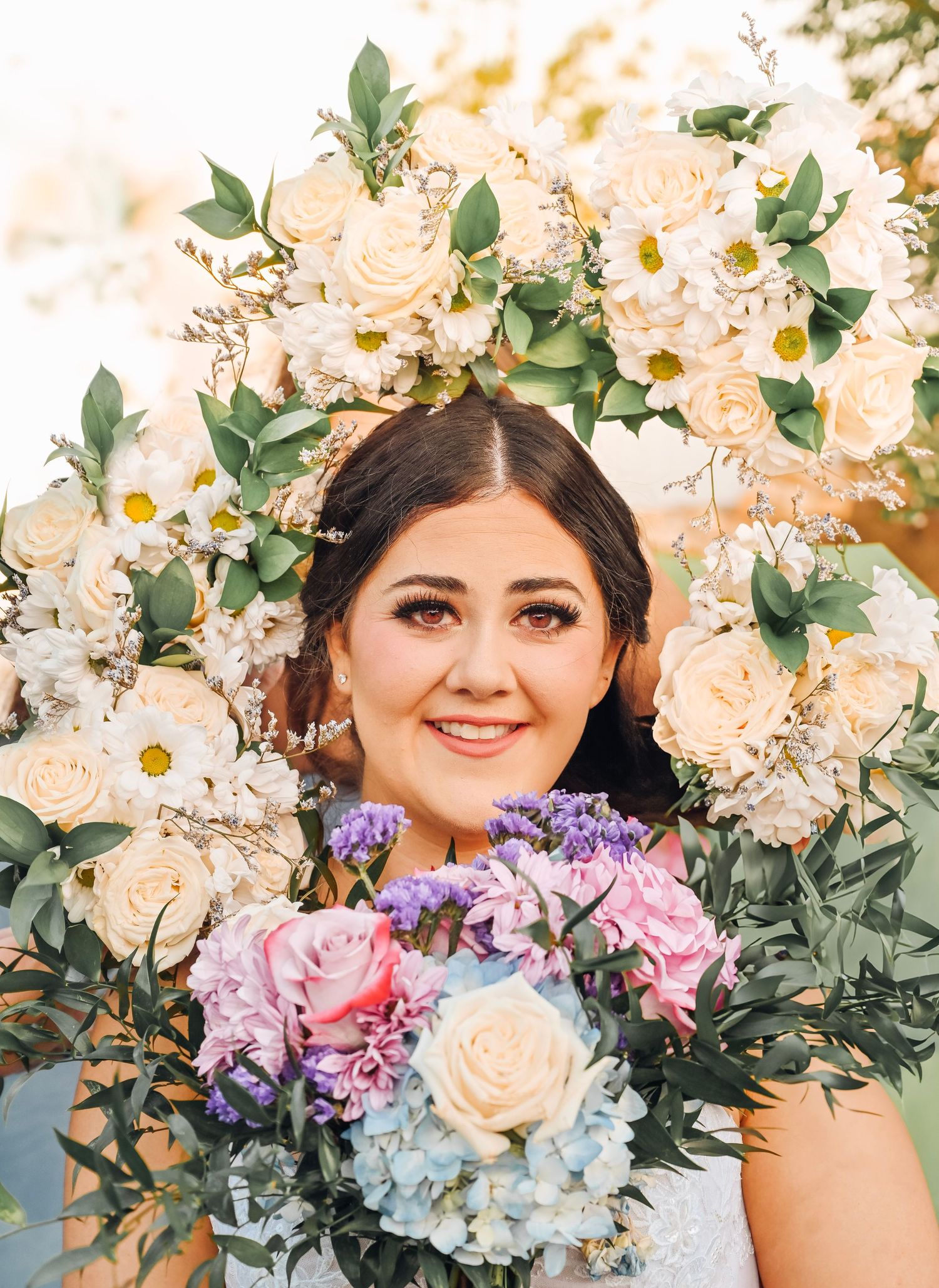 Bride smiling through a floral arch of white roses and greenery, holding a pastel bouquet of pink, purple, and blue flowers.