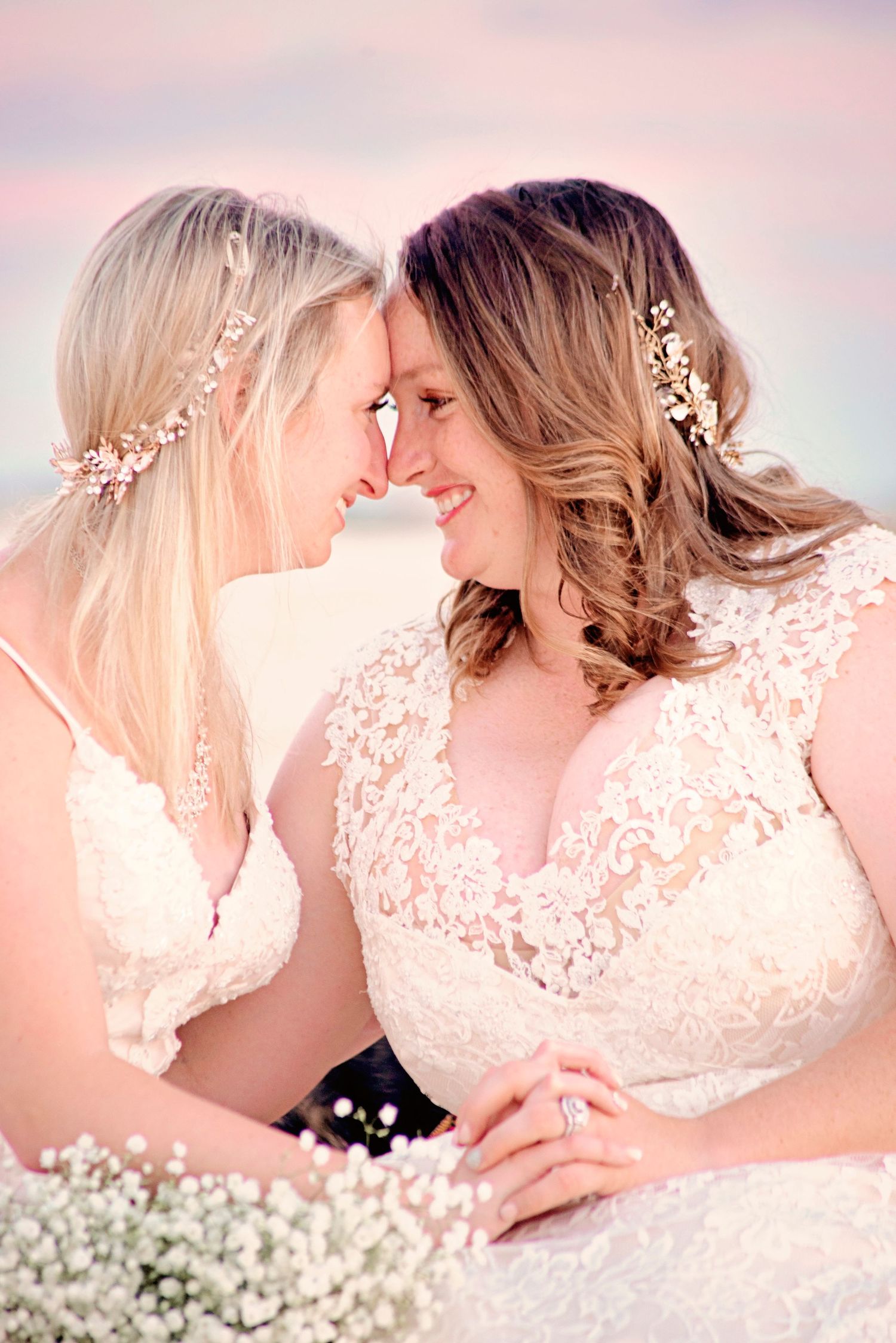 Two brides in lace wedding dresses touch foreheads romantically on a beach at sunset with baby's breath bouquet.