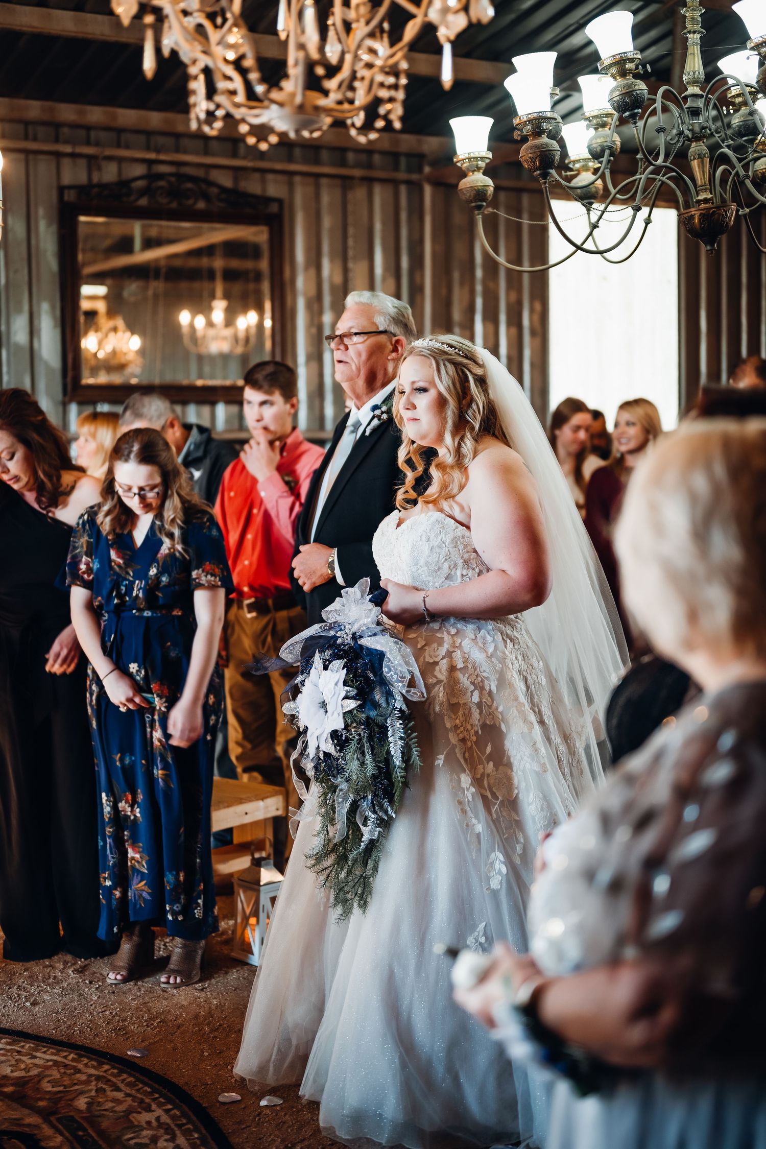 Bride in white gown and veil walks down aisle with escort in elegant chandelier-lit wedding venue.