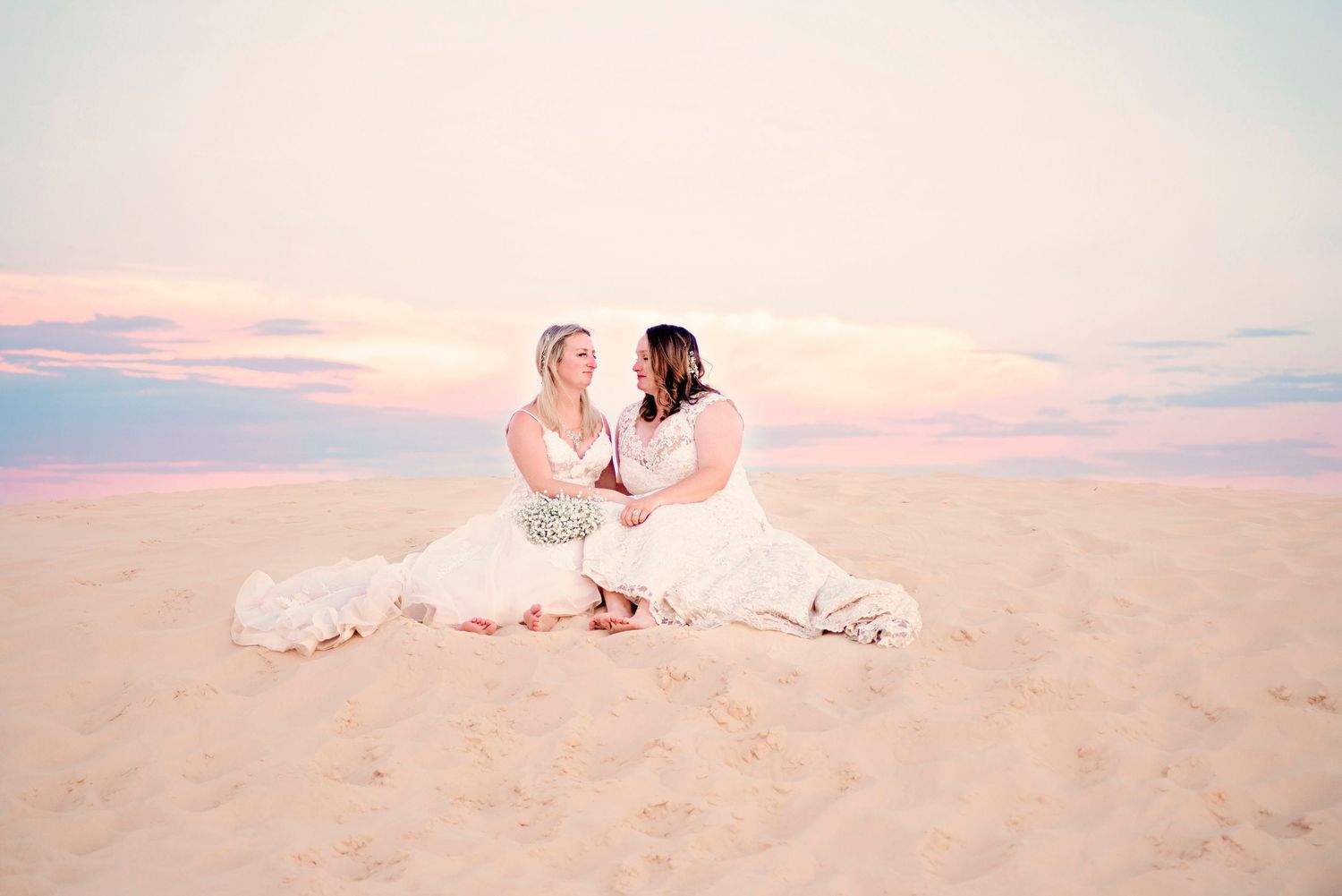 Two brides in white gowns sitting on sandy beach at sunset, facing each other romantically.