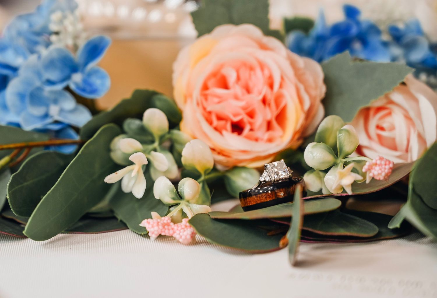 Wedding rings nestled among peach roses, blue hydrangeas, and eucalyptus greenery in an elegant floral arrangement.