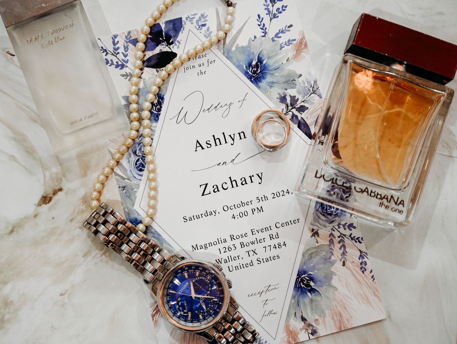 Wedding flat lay featuring an invitation for Ashlyn and Zachary, pearl necklace, watch, rings, and Dolce & Gabbana perfume.