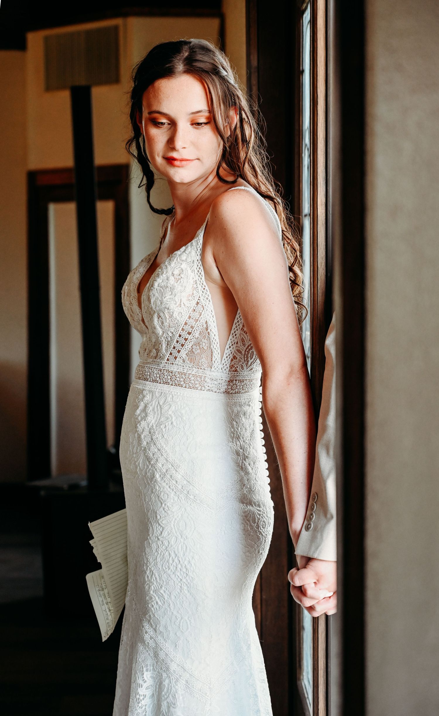 Brunette bride in fitted lace gown stands by window holding letter.