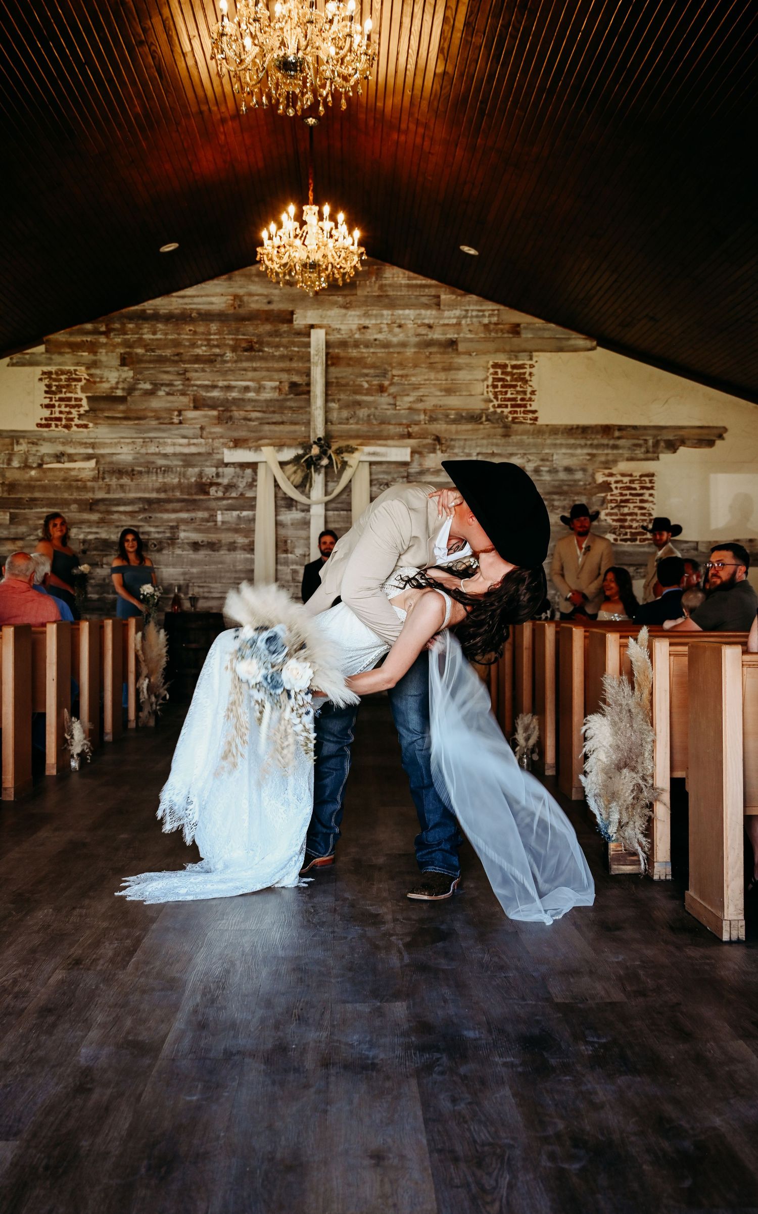 Groom dips bride for a kiss in rustic chapel aisle, chandeliers above, guests watching, western wedding style.