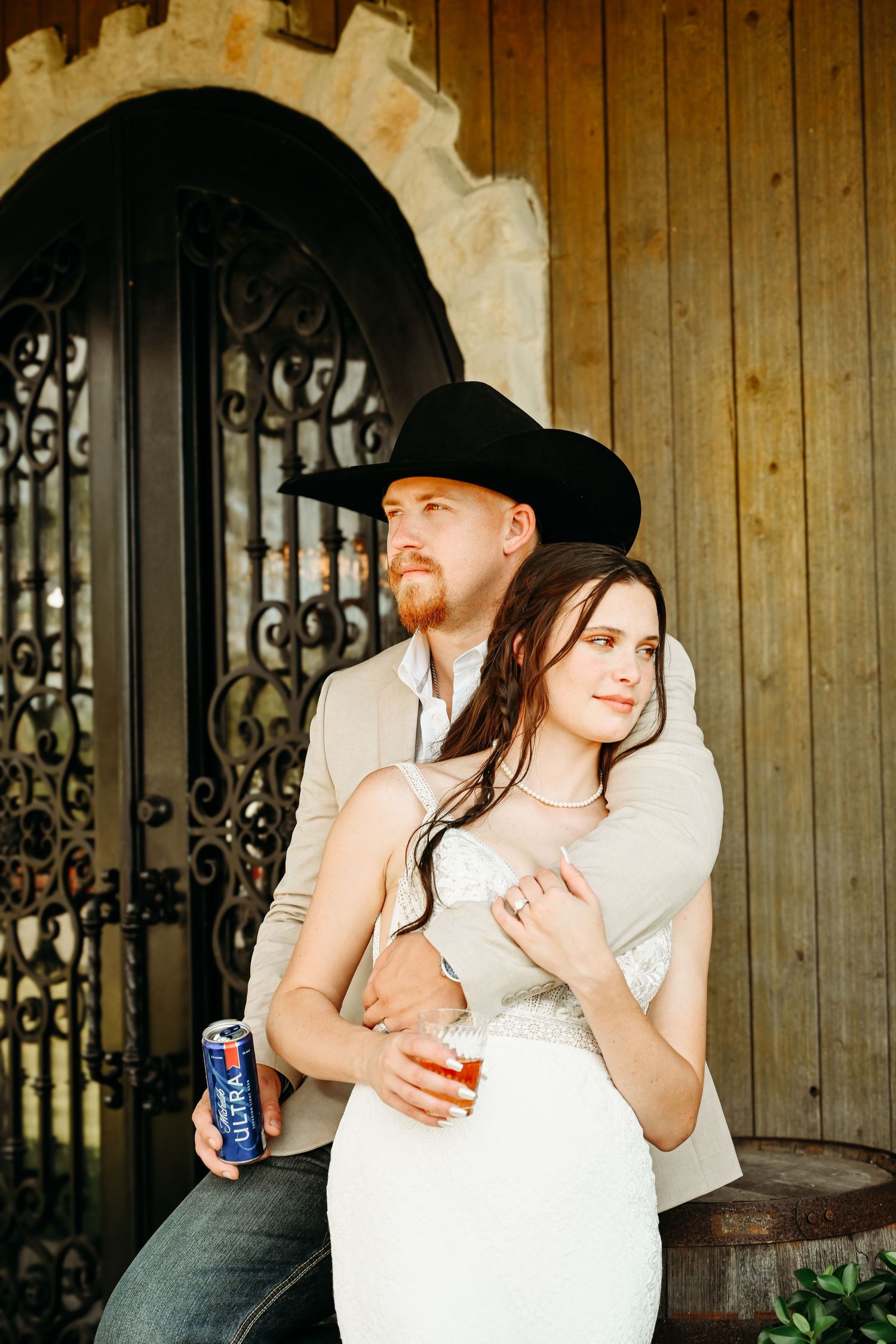 Couple in western wedding attire embrace outdoors, groom in black cowboy hat, bride holding a can of Michelob Ultra.