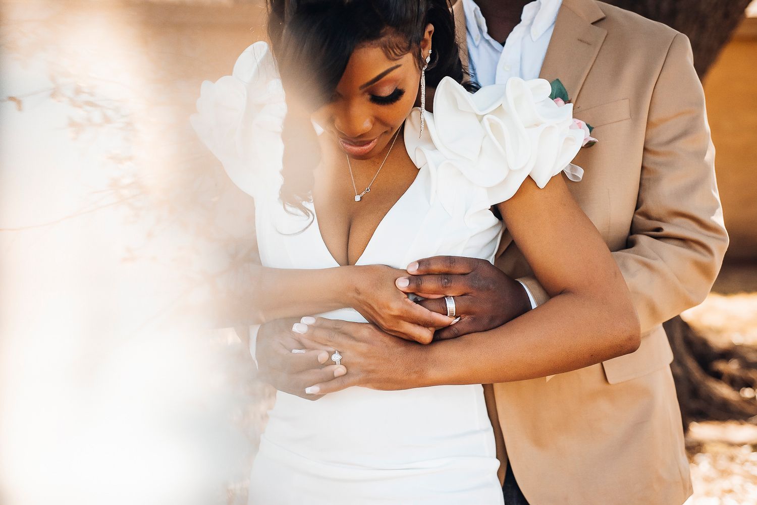 Bride and groom embracing tenderly, she in white gown, he in tan suit, exchanging rings outdoors.