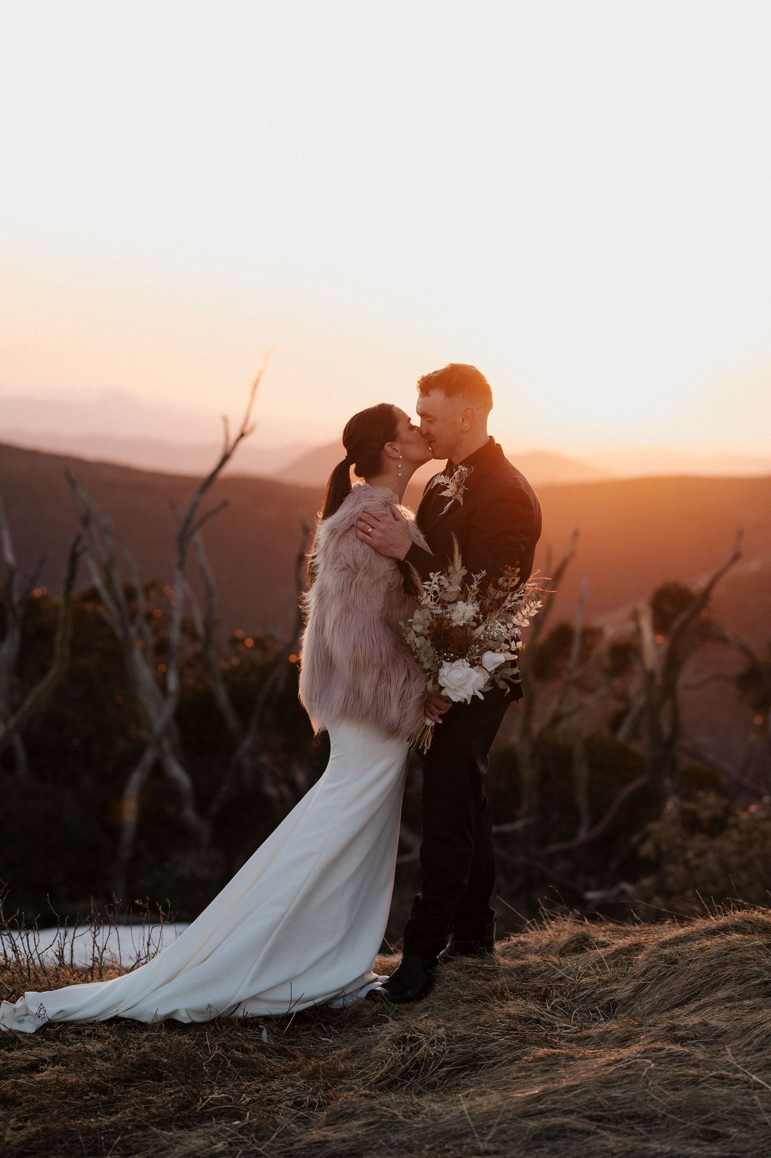 Romantic wedding couple sharing a moment at sunset on a mountaintop with white floral bouquet.