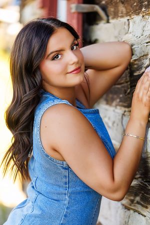 Young brunette woman in denim dress poses against rustic brick wall outdoors in warm sunlight.