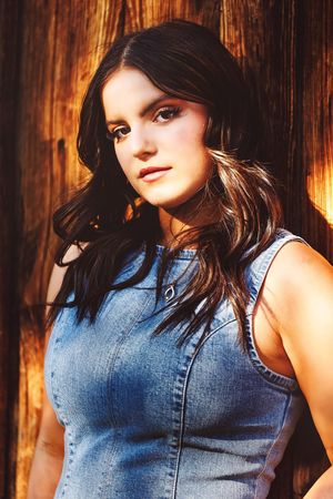 Young woman with dark hair wearing a denim sleeveless dress, posing against a warm wooden background.