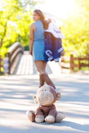 A worn monkey stuffed animal sits abandoned on a path as a young woman walks away in a sunny park.