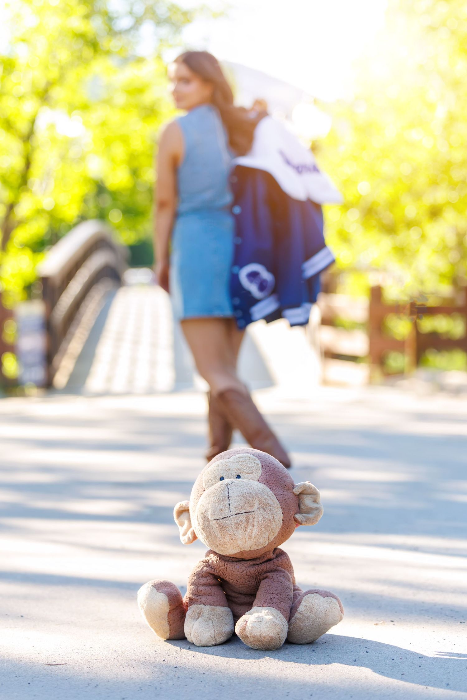 A worn monkey stuffed animal sits abandoned on a path as a young woman walks away in a sunny park.