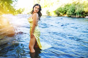 A woman in a yellow satin dress stands in a sunlit river surrounded by lush greenery and rocks.
