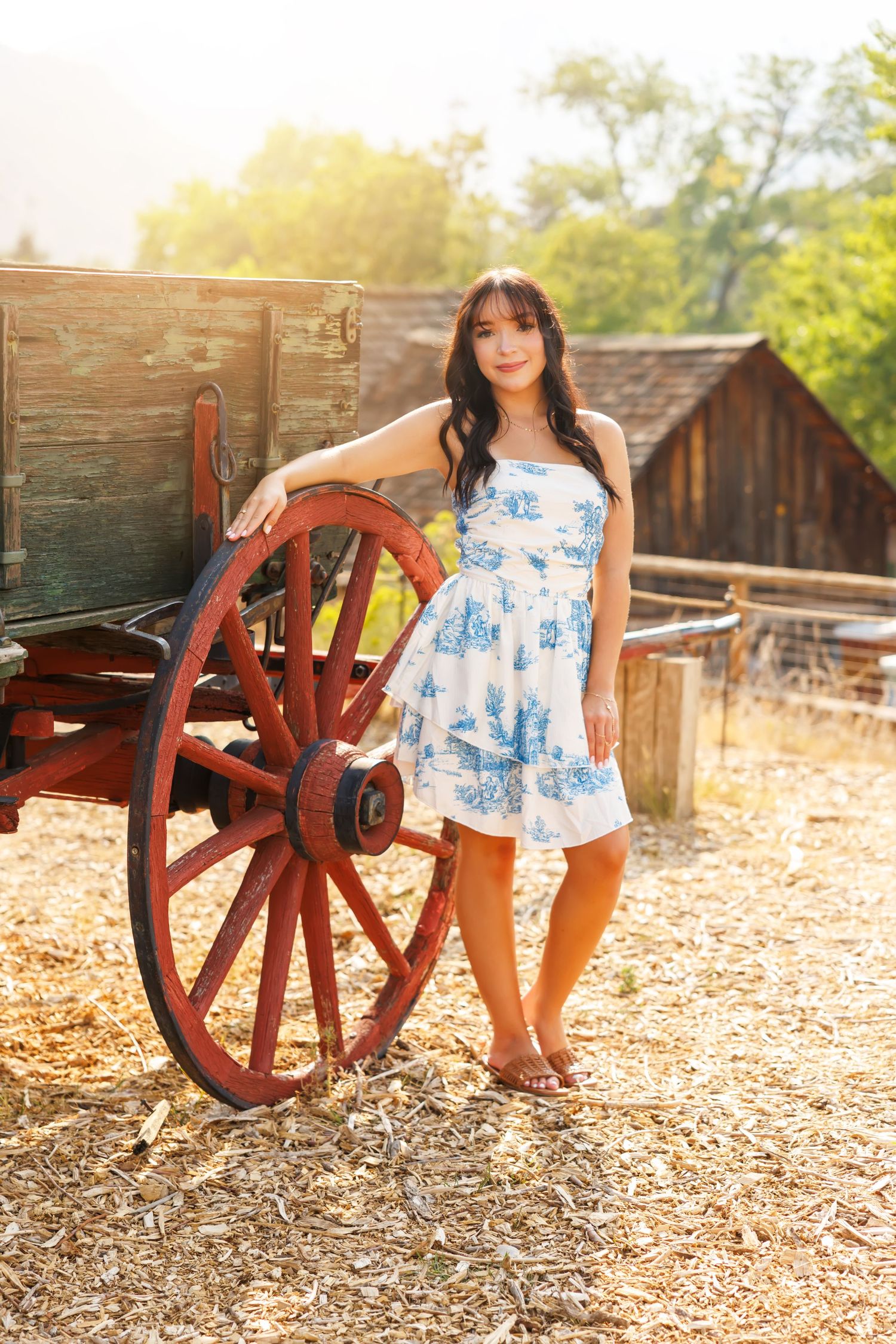 Young woman in blue floral sundress posing beside a vintage red wagon wheel on a sunny rural farm.