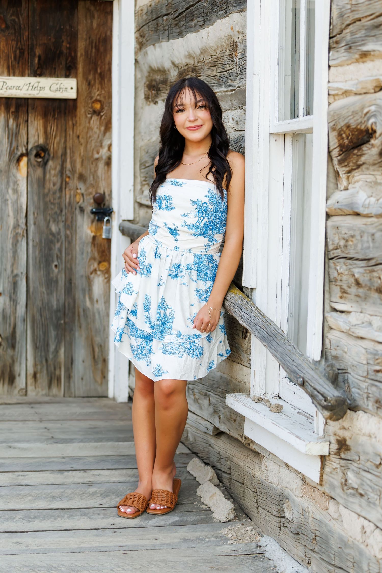 Young woman in blue floral sundress poses on rustic log cabin porch, smiling warmly in brown sandals.