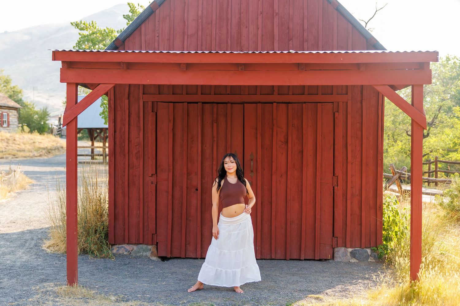 Woman in white skirt and brown top standing before a red barn in a sunny rural landscape.