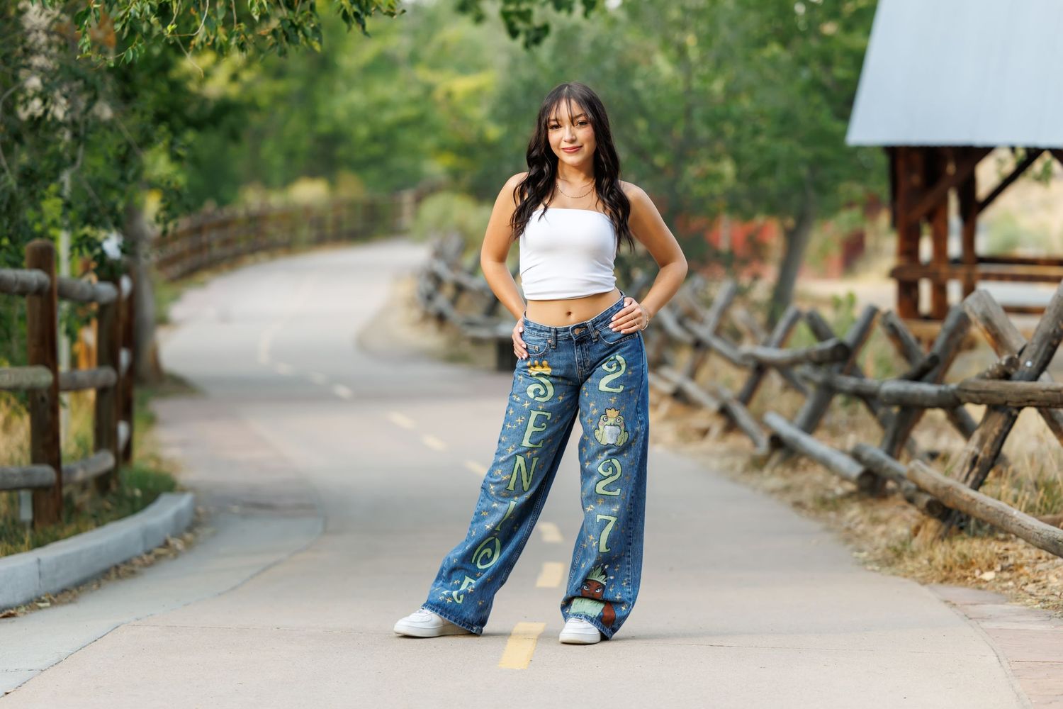 Young woman posing on a path in wide-leg graffiti jeans and white crop top, surrounded by lush greenery.