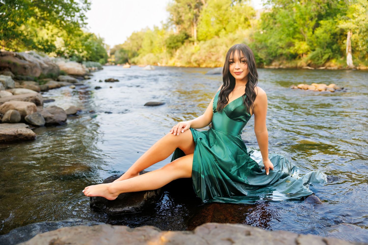 Young woman in elegant green satin dress sitting on rocks in a scenic river surrounded by lush greenery.