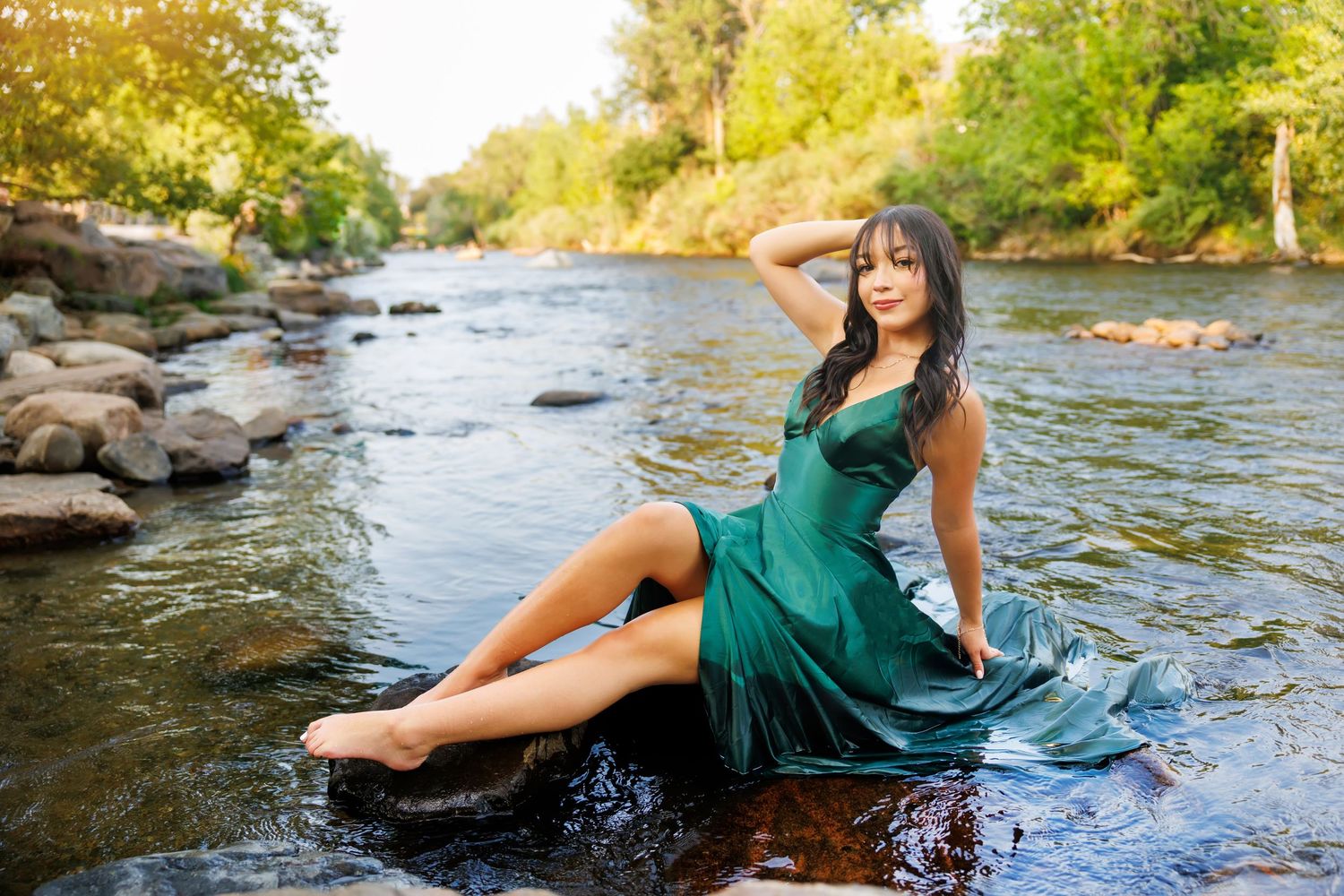 Woman in green dress sitting gracefully on rocks in a shallow river surrounded by lush trees.