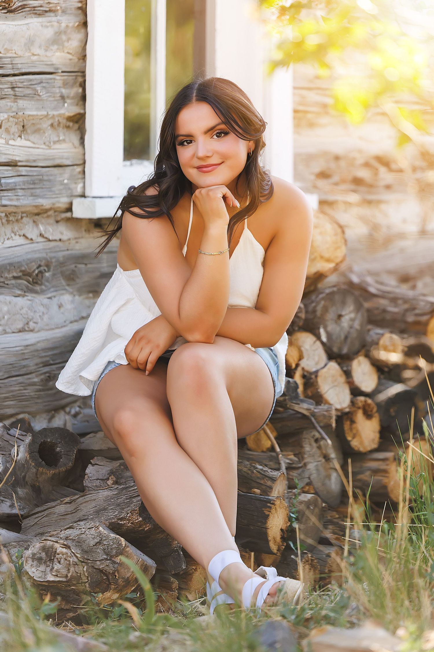 Young woman in white top and denim shorts sitting on logs outside a rustic cabin, smiling warmly in golden sunlight.