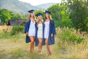 Three graduates in blue caps and gowns stand together in a sunny field with mountains visible in the background.